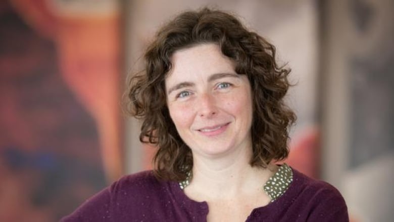 Close up portrait of a smiling woman with brown hair and wearing a plum coloured sweater with a blurry background