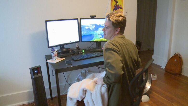 A man wrapped up in a blanket, near a heater, sits on a computer chair.