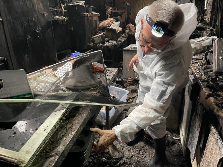 A man in disposable white protective coveralls and a headlamp sorts through an ash-covered room after a fire.