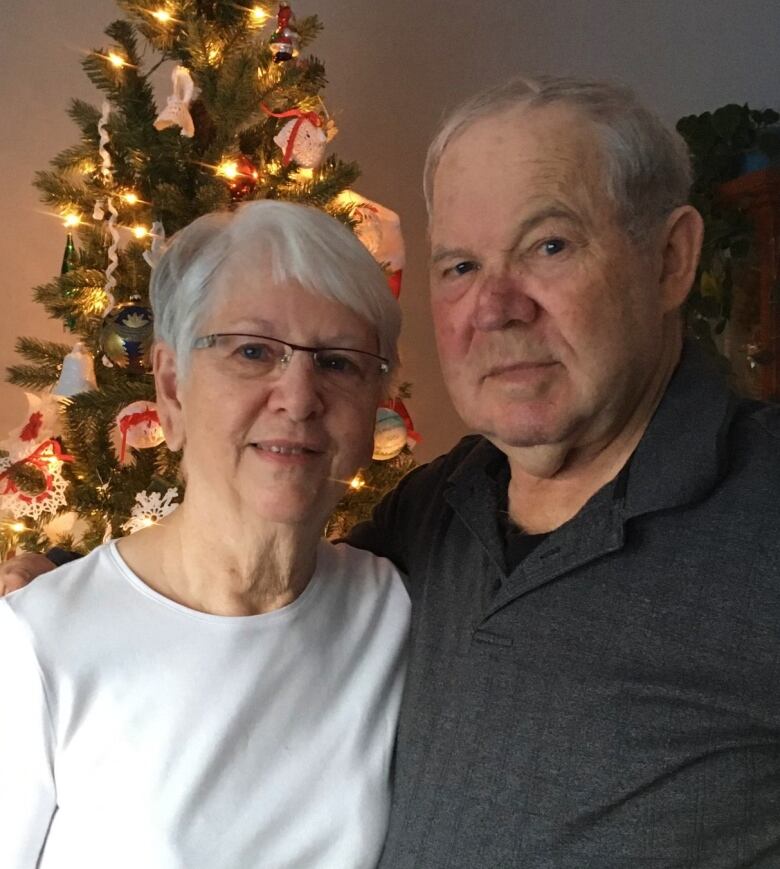 An older couple are smiling into the camera. They're standing in front of a Christmas tree.