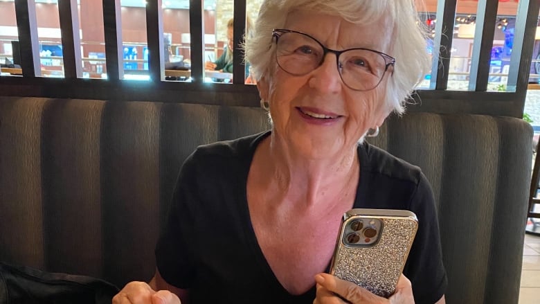 A smiling woman sits in a mall food court as she holds a phone.