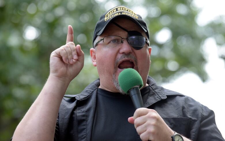 Man wearing a black cap, eye patch and black tee shirt speaks into a microphone