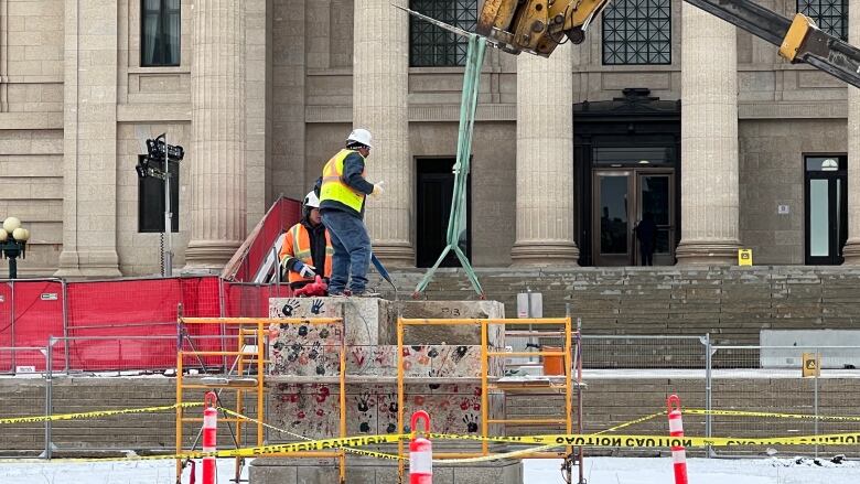 People in hardhats stand on top of a stone platform covered in handprints, while a piece of heavy machinery uses a rope to tie off the platform. Hundreds of orange flags cover the ground in front. 