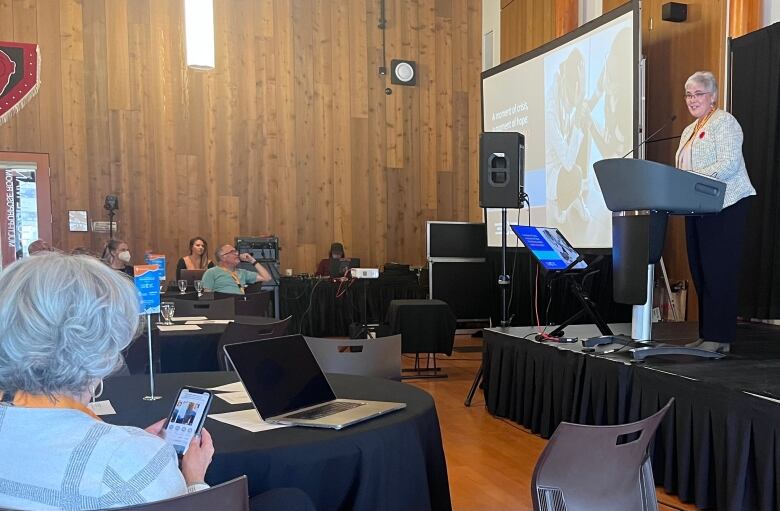 Dr. Kathleen Ross stands behind a lectern in a large conference room. People are sitting at tables watching as she presents a slide show.