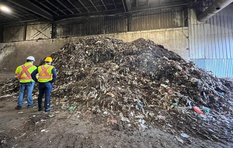 Two men in hard hats stand in front of a large pile of garbage.