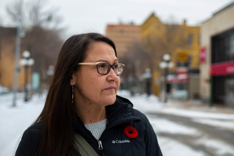 A woman wearing glasses in a winter jacket stands in a downtown area.