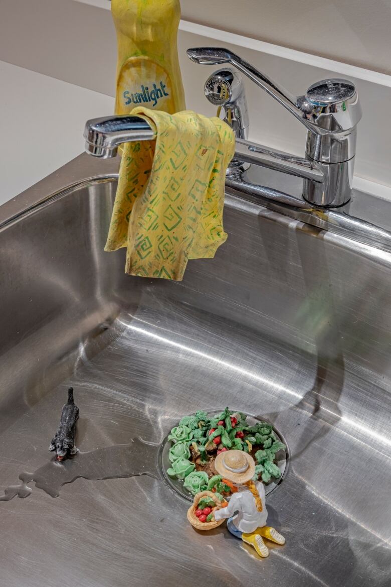 Photo of a kitchen sink. A miniature figure of a woman tends to a tiny clay vegetable garden in the drain.