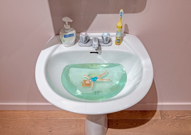 Top down photo of a bathroom sink filled with a sculpture resembling a tiny person floating in aqua-blue water.