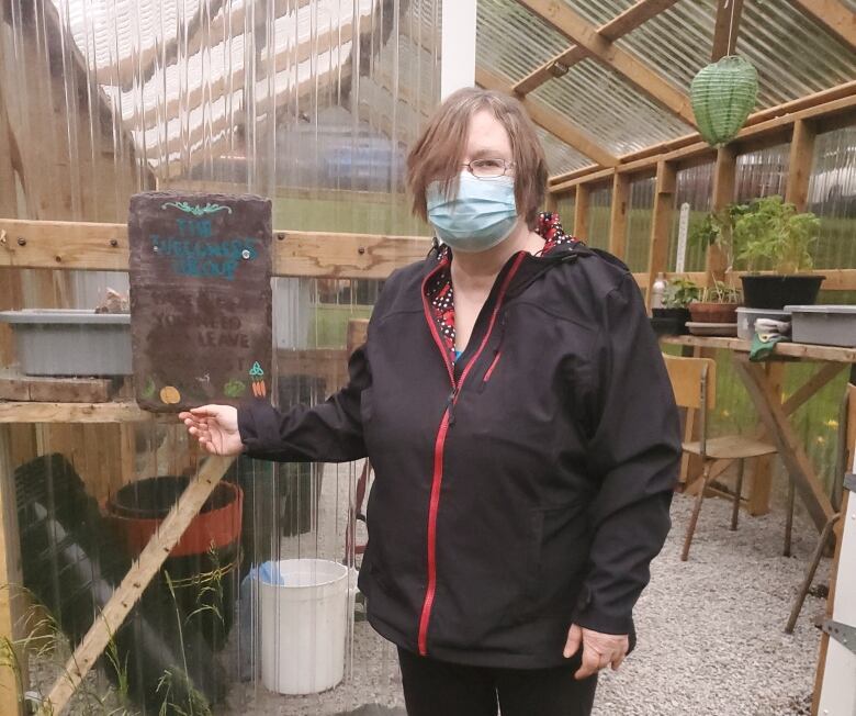 A woman wearing a black jacket and a mask poses in front of a glass greenhouse. She is pointing at the greenhouse sign she painted on a piece of 19th century slate.