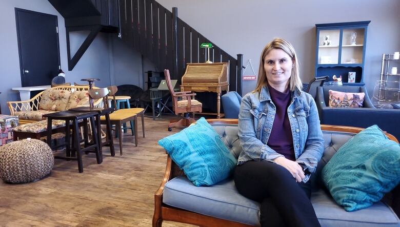 A woman wearing a jean jacket and a purple sweater sits on a small blue sofa inside a large open room. The room shows a variety of unusual pieces of furniture.