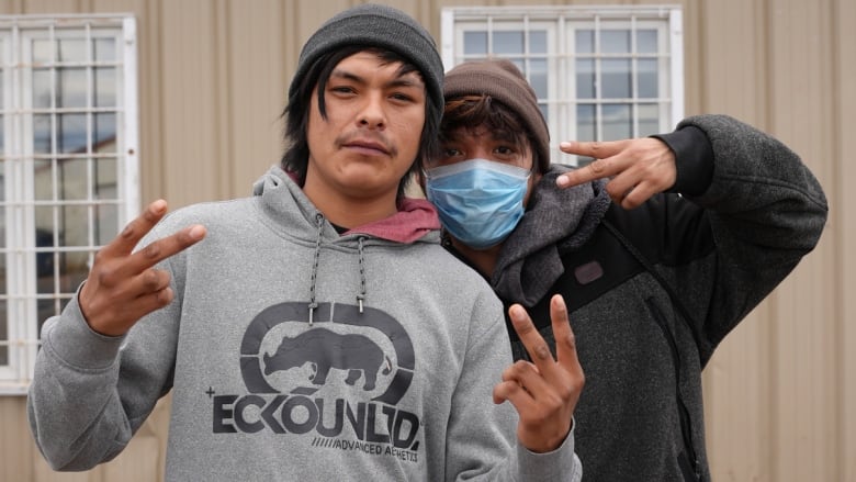 Two men in hoodies standing outside a beige building make peace signs with their fingers.