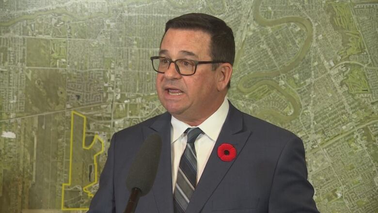 A man in a suit speaks into a microphone. There is a poppy on his lapel and he speaks in front of a large map background.