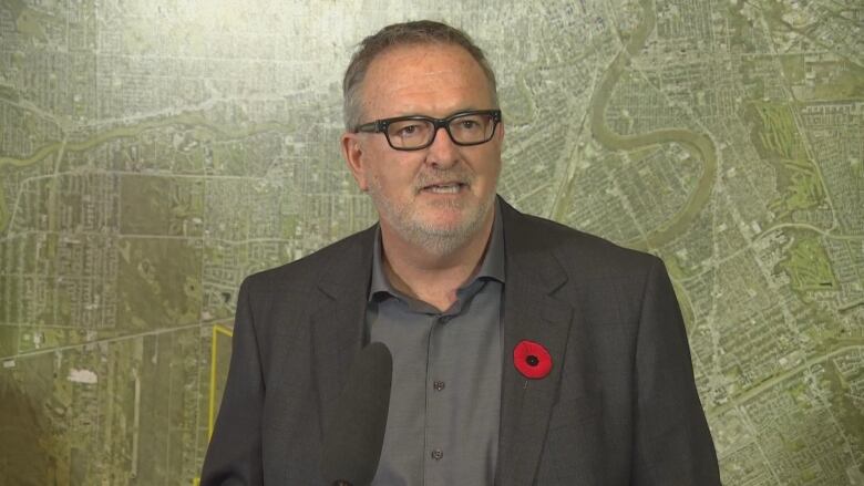 A man in a suit speaks into a microphone. There is a poppy on his lapel and he stands in front of a large map background.