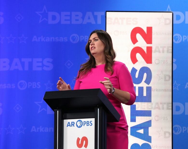 Individual with long brown hair wearing a dark pink dress, standing at a podium. 