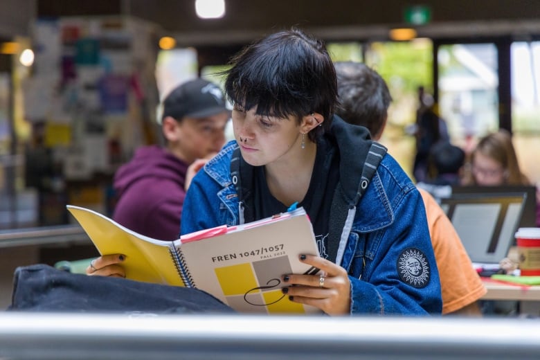 A student with short black hair and wearing a denim jacket reads through university course materials in a seated indoor area on campus, with other students seated and working behind them.