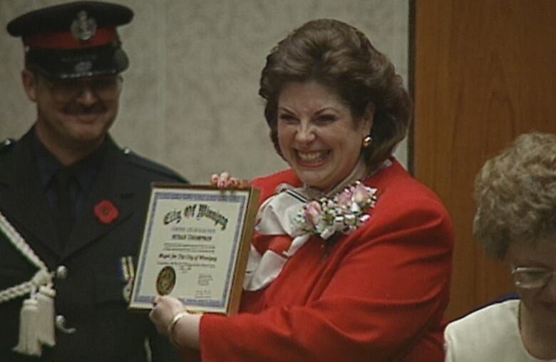 A woman with brown hair wearing a red suit, smiles broadly as she holds a cetificate declaring her mayor of Winnipeg, while a man wearing a police uniform smiles behind her.