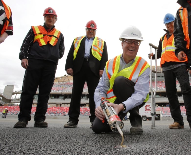 A man in a construction helmet and vest smiles while sealing a coin into the asphalt of a football stadium while other men in construction gear look on.