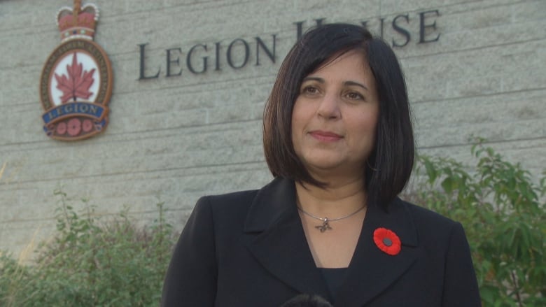 Woman with shoulder length hair standing in front of building wearing a blazer and poppy pin. Building has words 'legion house' written on it.