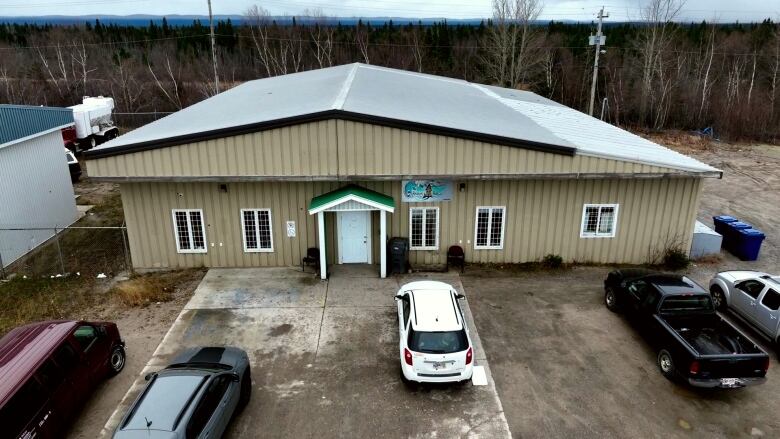 A square building with brown siding and a metal roof is shown from above. There are windows with bars on the front of it and a large wooded area behind. 