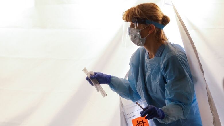 A nurse wearing a gown walks out a tent carrying a test kit for respiratory viruses.