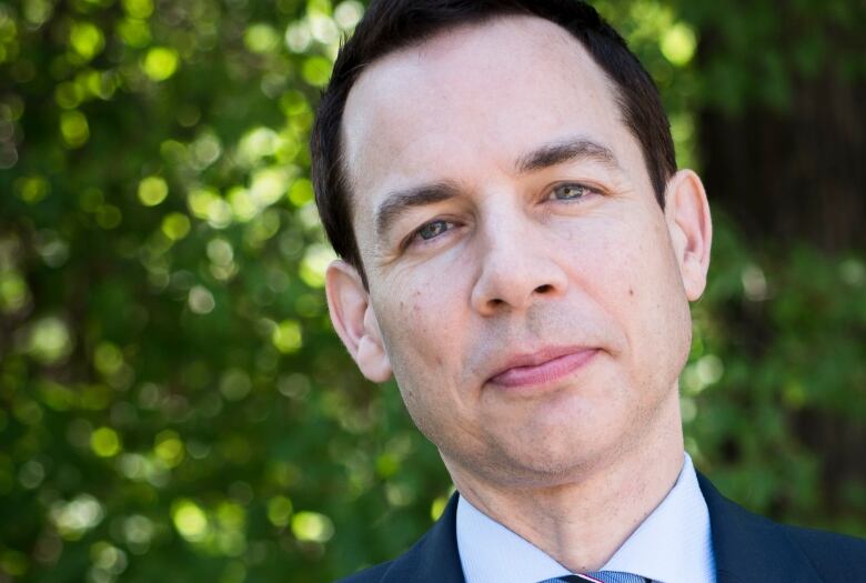 A man with dark hair and wearing a dark blue suit, light blue shirt and striped tie stands in front of leafy background.