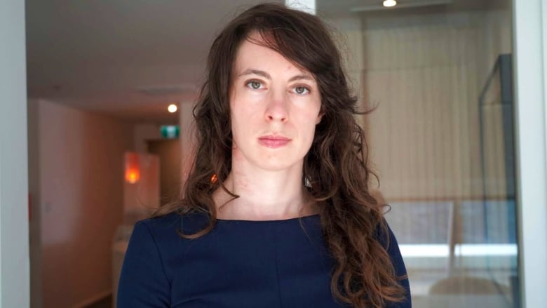 A white woman with long wavy brown hair looks directly at the camera. She is wearing a navy long-sleeved top and her facial expression is neutral.