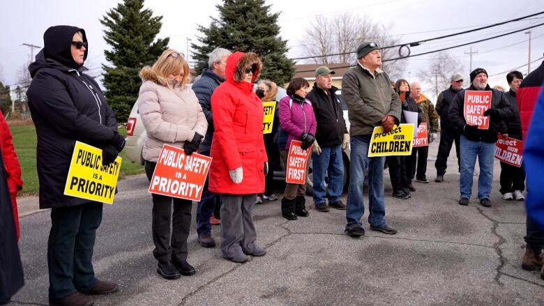 A group of people wear winter coats and hold signs saying 'public safety is a priority' and 'protect our children.'