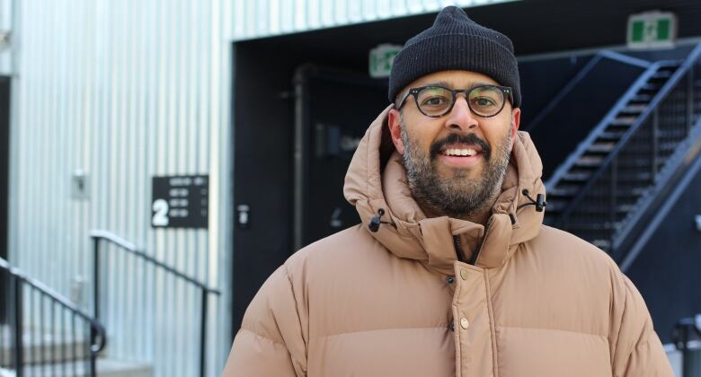 A smiling man with glasses and a black beard, wearing a black knit toque and tan winter jacket, stands in front of an industrial building.