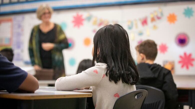 Students sit at their desks in a classroom with a teacher and a whiteboard in the background.