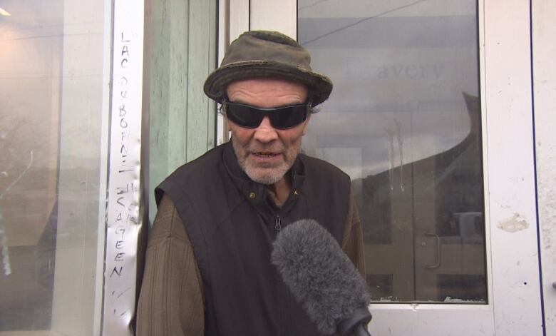 A man wearing a hat and sunglasses speaks into a microphone outside a door.