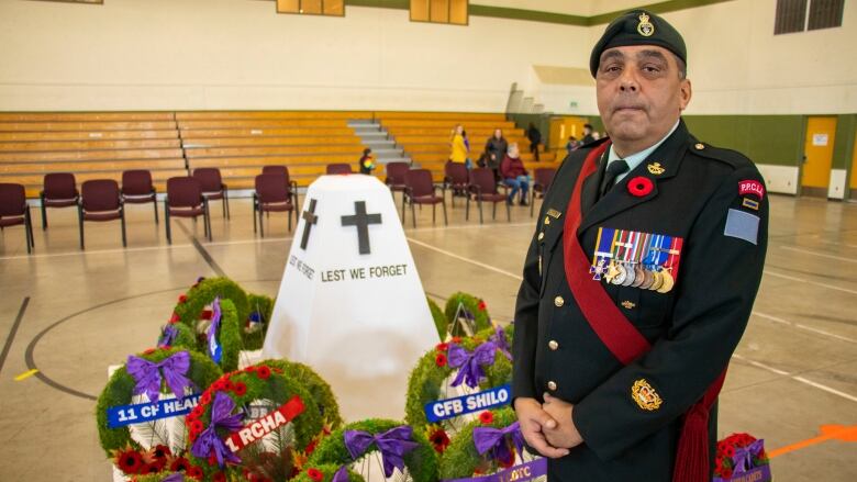 A man wearing a Canadian military uniform stands in front of a cenotaph in a gym space with poppy wreaths.