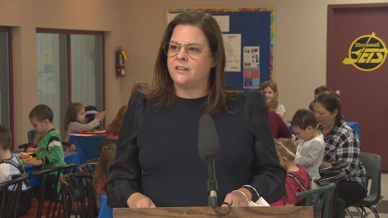 A woman with shoulder-length brown hair wearing aviator glasses and a black shirt stands at a podium in front of children playing with their caregivers.