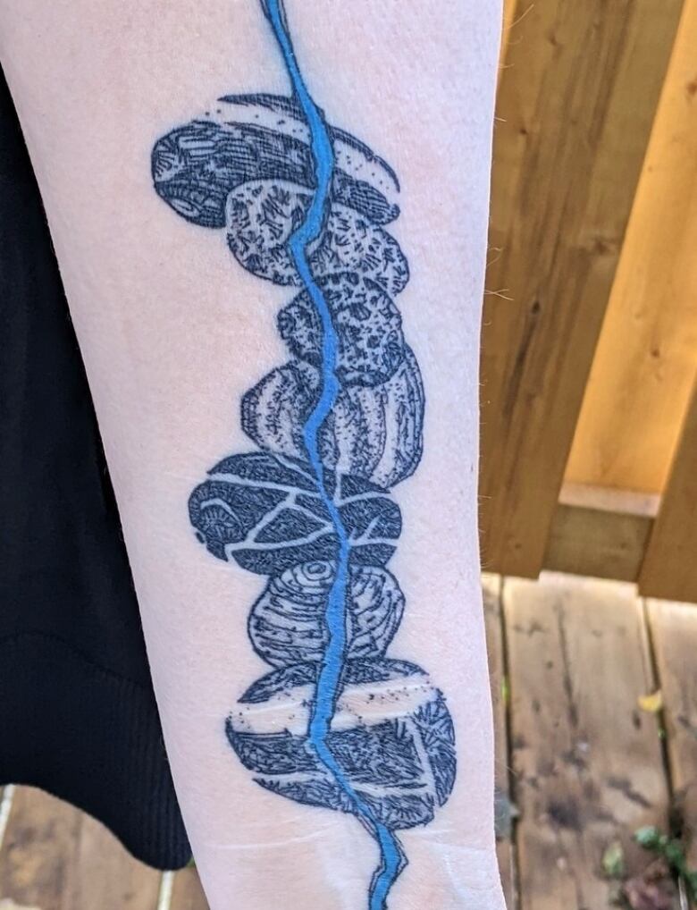 A photo of a tattoo on the inside of a person's arm. The tattoo shows rocks stacked on top of each other with a blue line going through the centre of them.