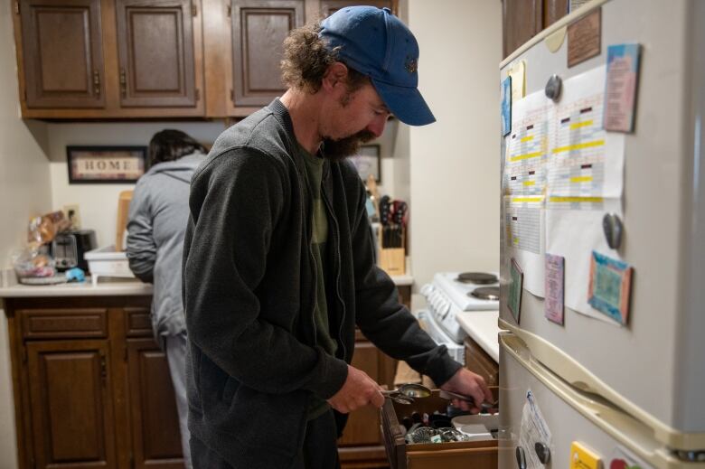 A man puts away dishes in the foreground of the photo, while a woman behind him stands with her back to the camera at the kitchen sink.