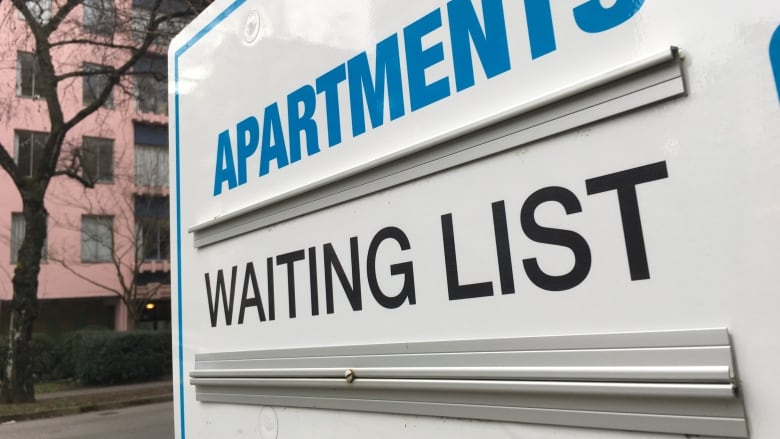 A no vacancy sign with the word Apartments in blue and the words Waiting List in black on a white board in front of a pink brick highrise building.