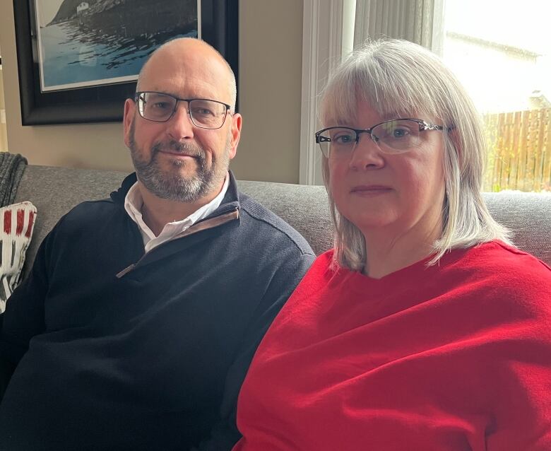 A man, left, and a woman, both middle-aged, sit next to each other on a couch and look into the camera.