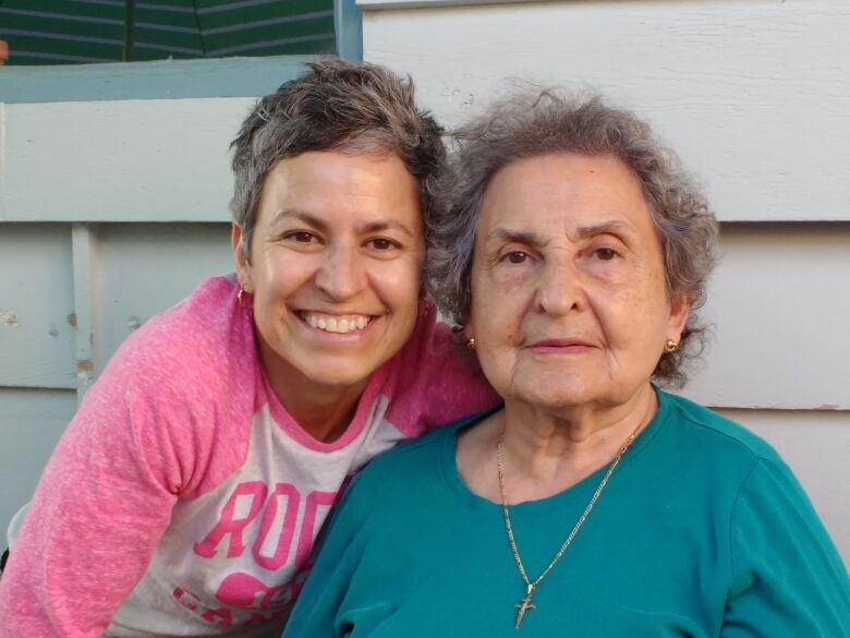 A smiling middle-aged woman with short salt-and-pepper hair crouches next to an older woman wearing a turquoise shirt with a gold cross necklace.