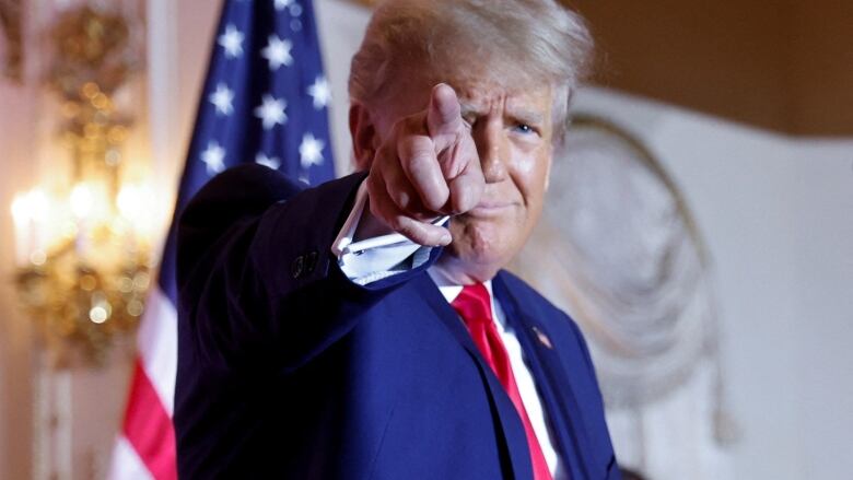 Man with blue jacket and red tie gestures #1 towards the camera from a stage with American flags behind him. 