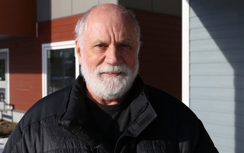 A balding man with white hair and a beard wears a black winter jacket.