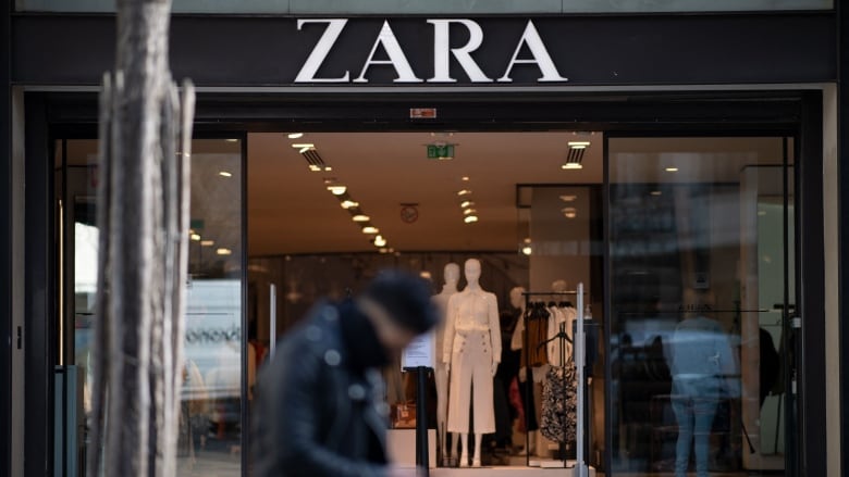 A man sits on a bench in front of a clothing store with fashion mannequins in the window. 