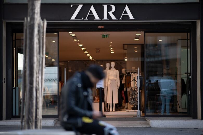 A man sits on a bench in front of a clothing store with fashion mannequins in the window. 