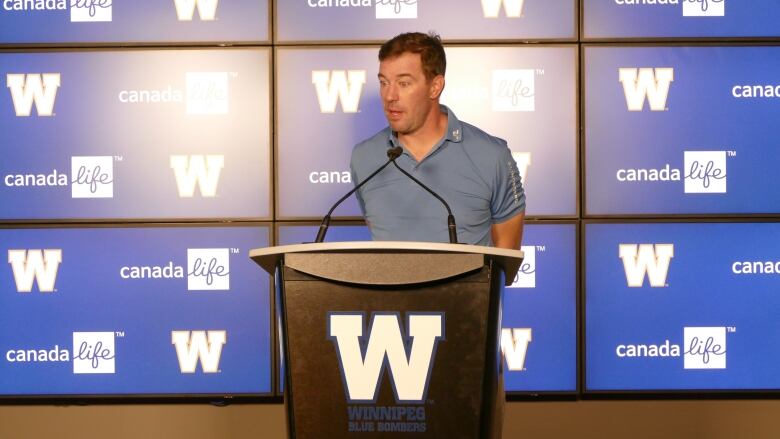 A man speaks at a podium with screens displaying the Winnipeg Blue Bombers logo behind him.