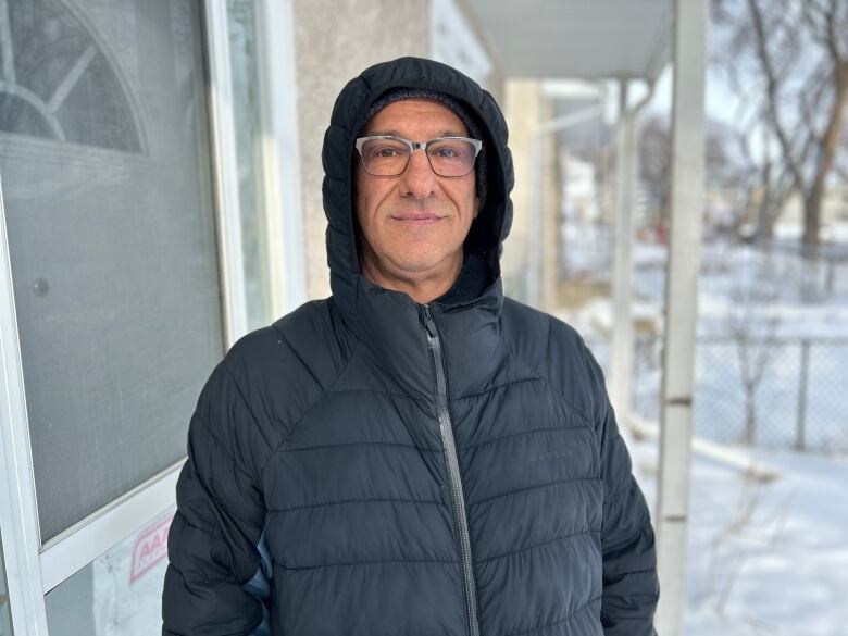 A man wearing a winter jacket with the hood pulled up is standing on a porch in front of a house and smiling at the camera. 