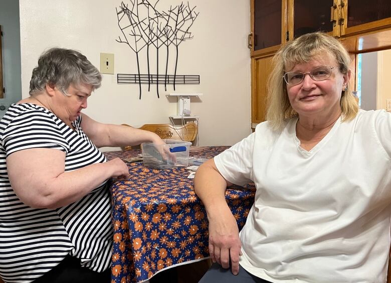 Two middle-aged women sit at a table. The one on the left is reaching into a box on the table and is sitting sideways to the camera. The other one, on the right, is turned towards the camera.