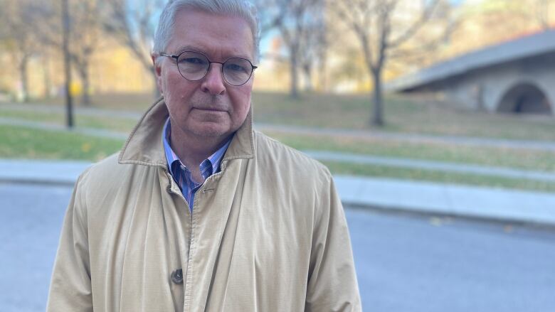 A man in a beige trench coat poses for a photo.