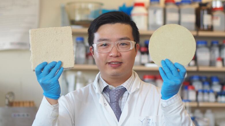 University of British Columbia postdoctoral fellow Dr. Yeling Zhu wears a white lab coat, blue latex gloves and safety goggles. He is photographed from the chest up and, in each hand, holds a block of foam.