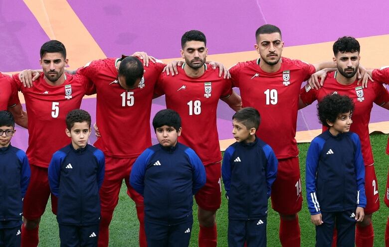 Men wearing red jerseys stand shoulder to shoulder, arms links, with solemn faces, and children wearing blue jerseys stand in front of them. 