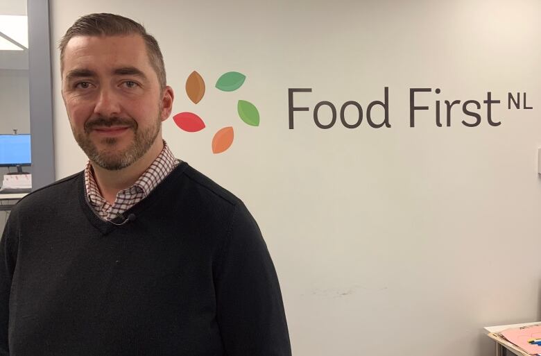 A person in a black sweater stands in front of a wall with the Food First NL logo.