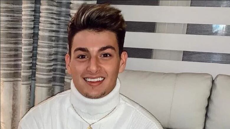 A young man wearing a white turtle neck smiles at camera.