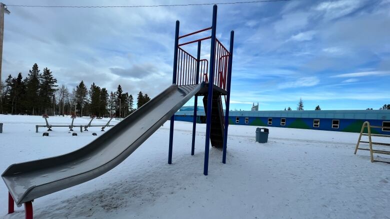 A school yard slide is seen in front of a low-lying elementary school painted in two tones of blue.